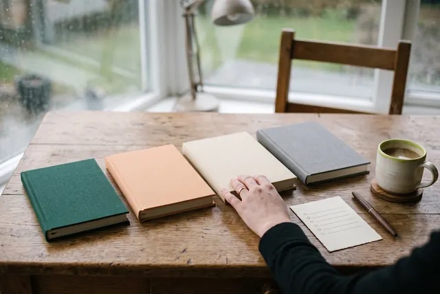 Four closed notebooks in different colours side by side on a desk, a hand resting on one