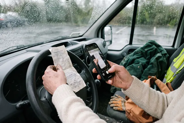 A tradesperson in a parked van photographs a crumpled till receipt with a phone