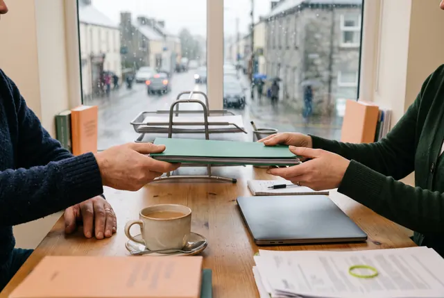 A sole trader hands a green folder of records across a desk to an accountant